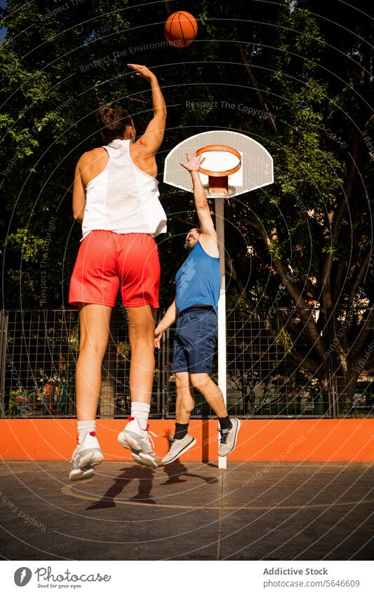 Full length back view of sportswoman in activewear shooting basketball towards basket and man trying to defend on court against sky Sportspeople Basketball