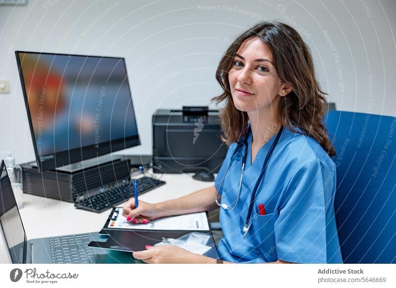 Young woman doctor sitting at table with laptop smartphone and making note on clipboard write smile stethoscope pen clinic female young professional specialist