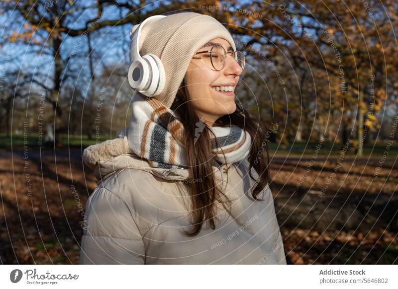 Smiling woman in headphones listening to music in autumn park smile song positive enjoy leisure warm clothes glasses wireless female young happy eyeglasses hat