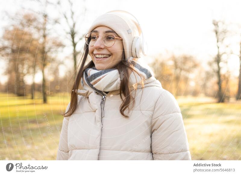 Smiling woman in headphones listening to music in autumn park smile song positive enjoy leisure warm clothes glasses wireless female young happy eyeglasses hat