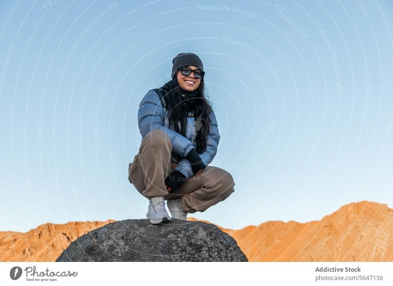 Happy woman hiker looking at camera while sitting on rock in warm clothes and sunglasses in daylight traveler tourist smile mountain blue sky explore nature