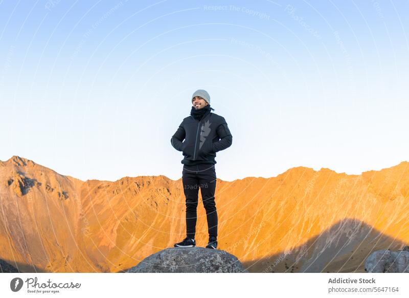 Full body of Happy man hiker standing on rock with hands in pockets in daylight traveler tourist smile mountain blue sky explore nature male happy cheerful