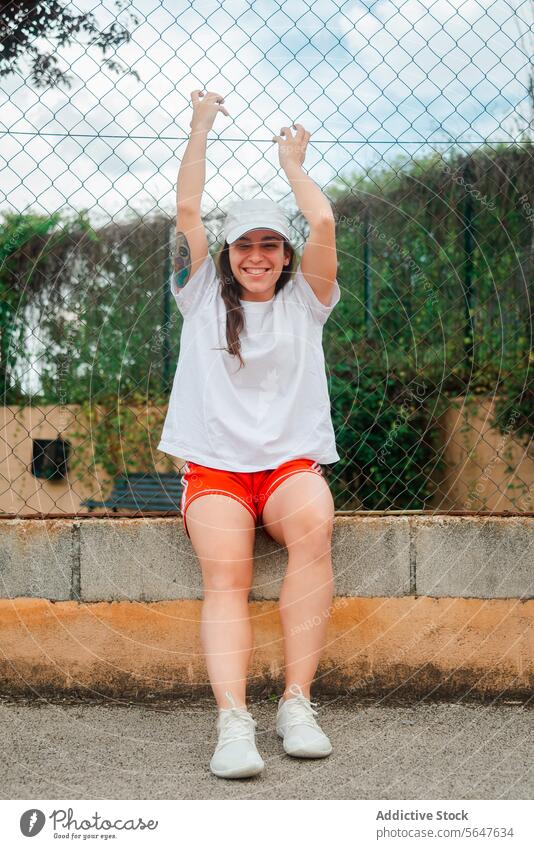 Cheerful young woman during workout break outdoors joy happiness sporty attire sitting curb foliage background cheerful casual clothing white shirt red shorts