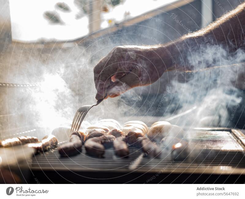 Unrecognizable man grilling sausages on grill rack covered by smoke chef cook poke fork prepare barbecue kitchen male adult hand stand meat process mushroom