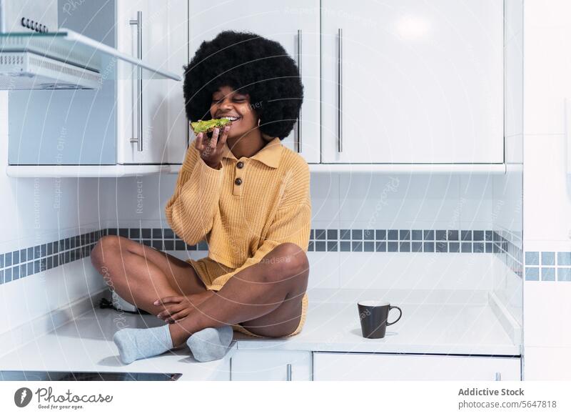 Cheerful black woman sitting at kitchen breakfast morning avocado home relax eat counter enjoy female african american afro curly hair food hot drink toast