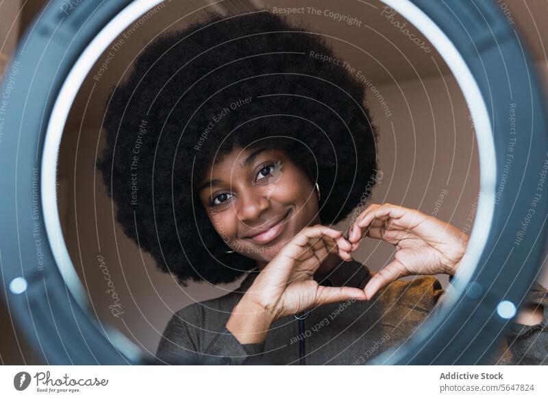 Smiling black woman showing heart gesture through ring lamp blogger love vlogger leisure smile afro hairstyle feminine appearance happy charming brunette