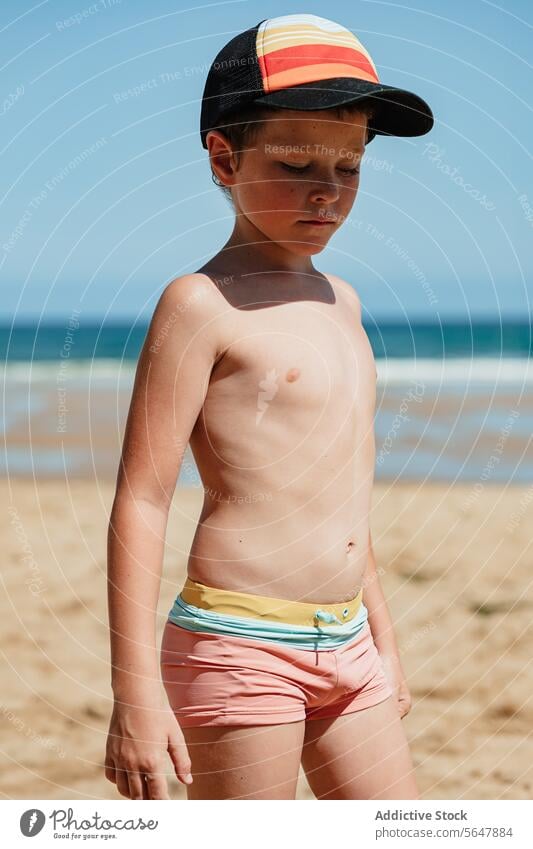 Thoughtful young boy on the beach with baseball cap child contemplative thoughtful sand ocean blue sky swimwear summer sea coastline leisure vacation holiday