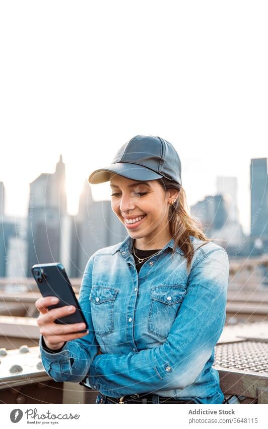 Woman using smartphone with Manhattan backdrop woman sunglasses cap smile skyline cityscape New York outdoor urban bridge fashion leisure casual denim sunset