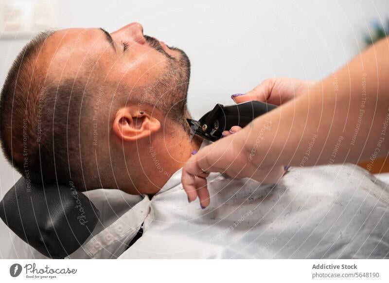 Side view of cropped unrecognizable female barber's hands shaving closed eye male client's beard with a trimmer machine at salon Hairdresser Shave Beard Man