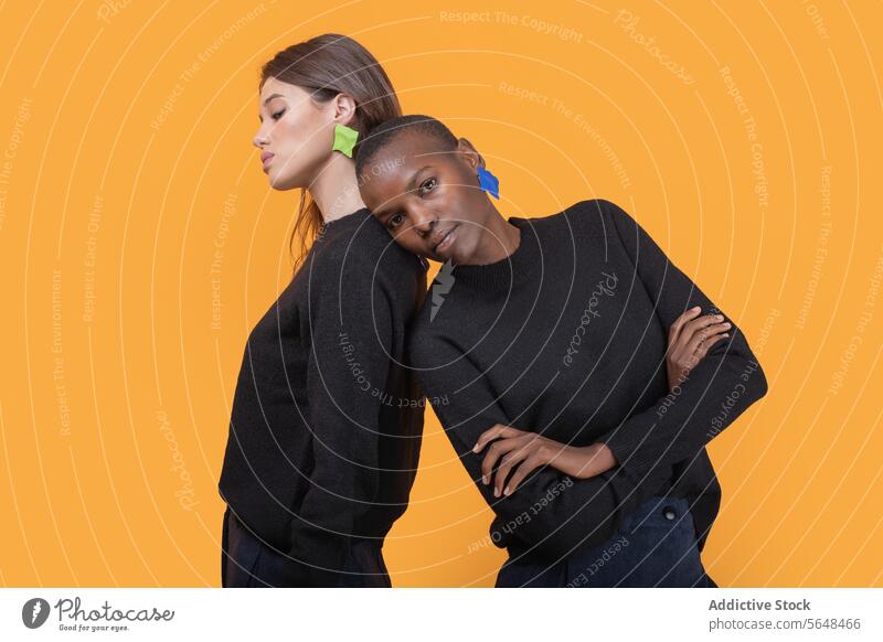 Side view of diverse young female friends leaning on each other looking at camera while wearing trendy colorful earrings against yellow background women