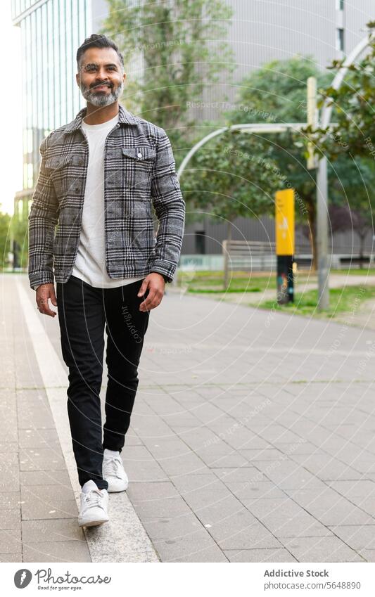 Cheerful Indian businessman in casual outfit walking along street cheerful indian stroll smile happy daytime male pavement city sidewalk positive optimist urban