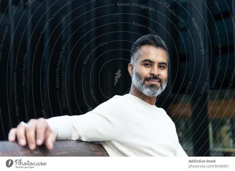 Happy Indian businessman sitting on wooden bench and looking at camera casual outfit beard indian alone lean on park entrepreneur male urban confident daytime