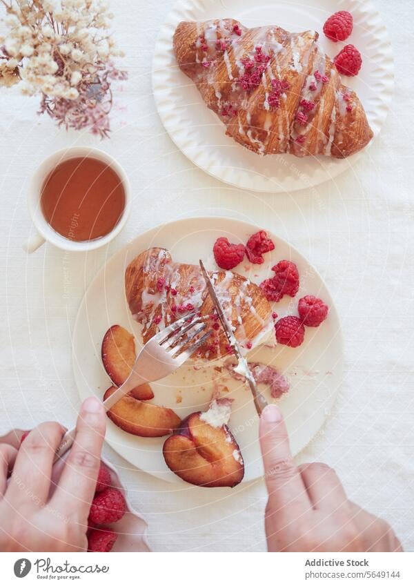 Crop person cutting croissant at restaurant hand raspberry peach knife cup breakfast dessert fork plate ceramic tea tasty delicious table fresh beverage sweet