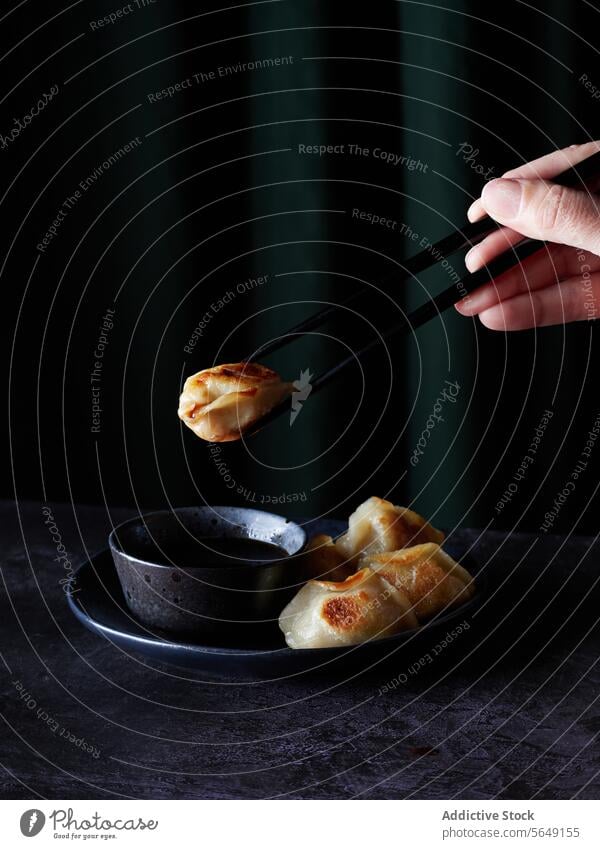 Anonymous person holding chopsticks that delicately grip a golden dumpling against dark green background dumplings sauce hand sensory experience slate plate