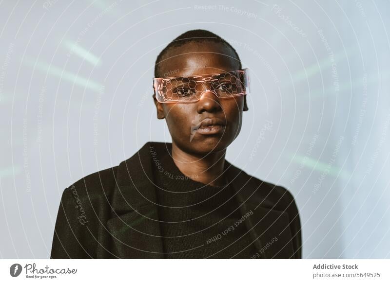 Portrait of confident young bold African American woman in smart futuristic VR glasses and long black trench coat standing looking at camera against neon light illuminated background