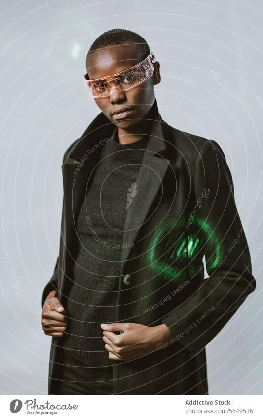 Portrait of confident young bold African American woman in smart futuristic VR glasses and long black trench coat standing looking at camera against neon light illuminated background