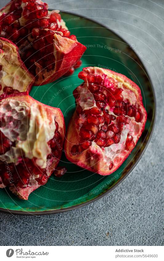 Fresh pomegranate pieces with vibrant red seeds on a green ceramic plate set against a textured grey concrete background fruit juicy ripe open section food