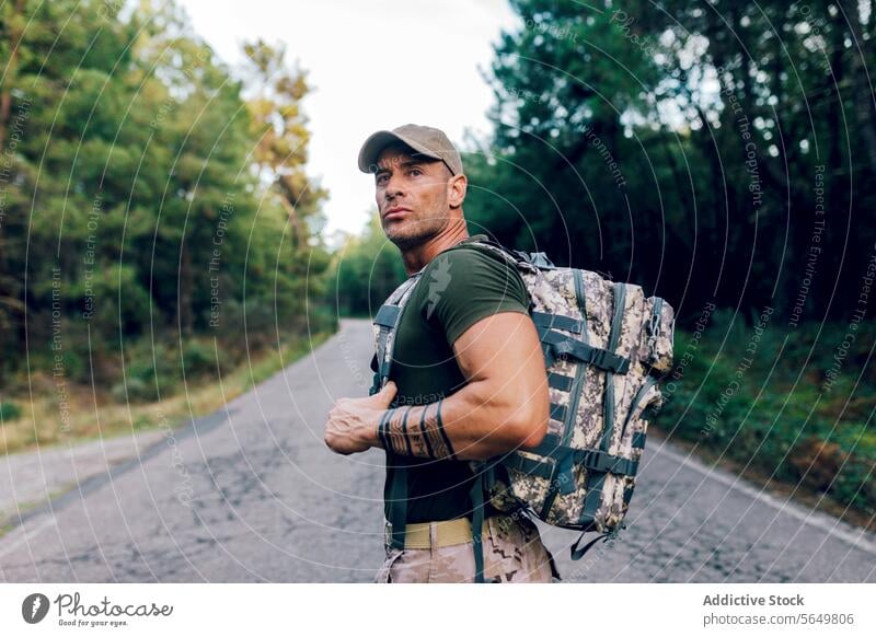 Army man in uniform with backpack and cap standing on road against trees in forest soldier confident camouflage pant green looking away army military warrior