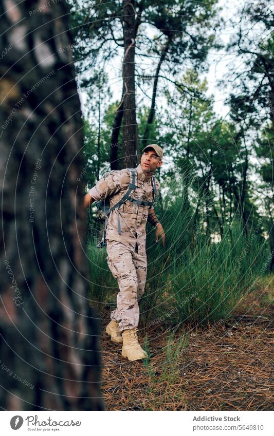 Full body of Army commando with cap and backpack standing in forest and looking away soldier military camouflage uniform blurred background army special force