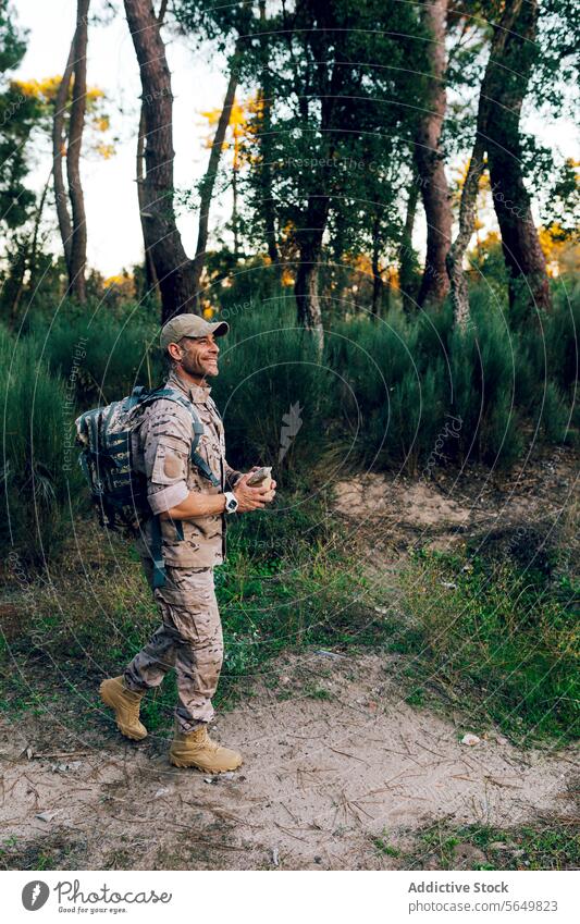 Full body of Army commando with cap and backpack holding box standing in forest and looking away soldier military camouflage uniform blurred background army