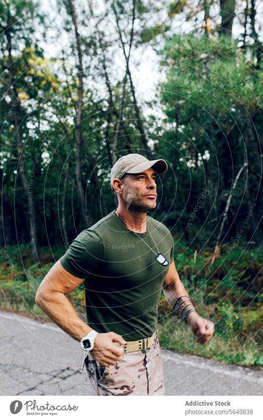 Confident mature male commando running in nature while training in forest soldier military jogging fitness road tree routine confident camouflage green pant cap