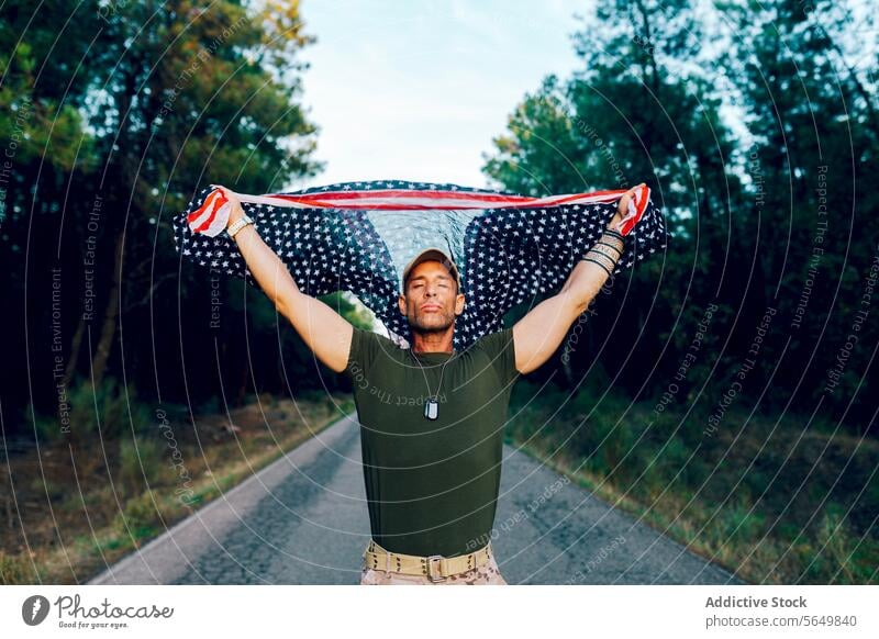 Military soldier with closed eyes waving national flag with pride while standing on road against forest man commando american aloft uniform special force