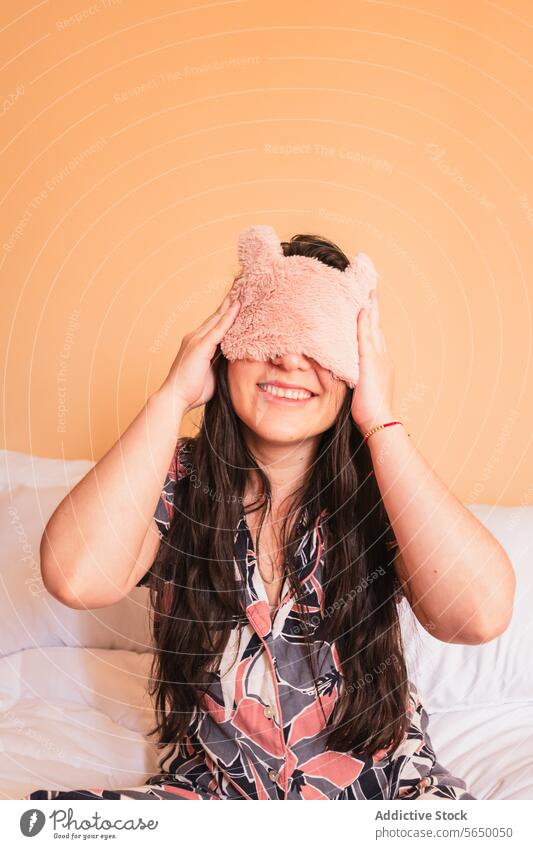 Smiling woman with sleep mask sitting on bed smile pajama cover eyes sleepwear comfort cozy bedroom female young cheerful nightwear joy awake happy glad