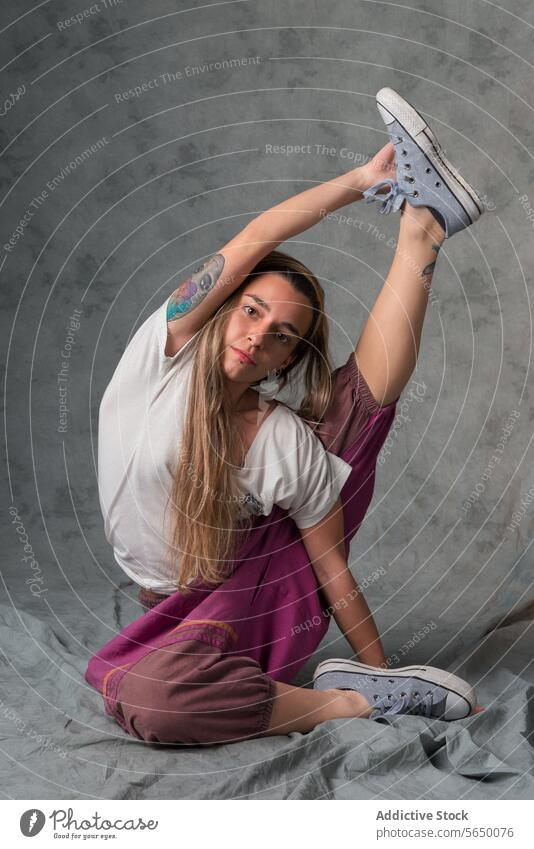 Full body of young woman in flexible yoga pose, stretching her leg upwards looking at camera Yoga flexibility casual wear fitness health wellness fabric seated