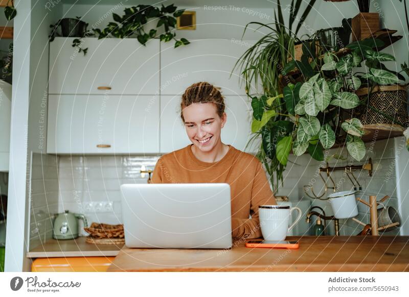 Happy Young woman working on laptop at kitchen table with smartphone and coffee mug in daylight cabinet at home female young dreadlocks gadget device mobile