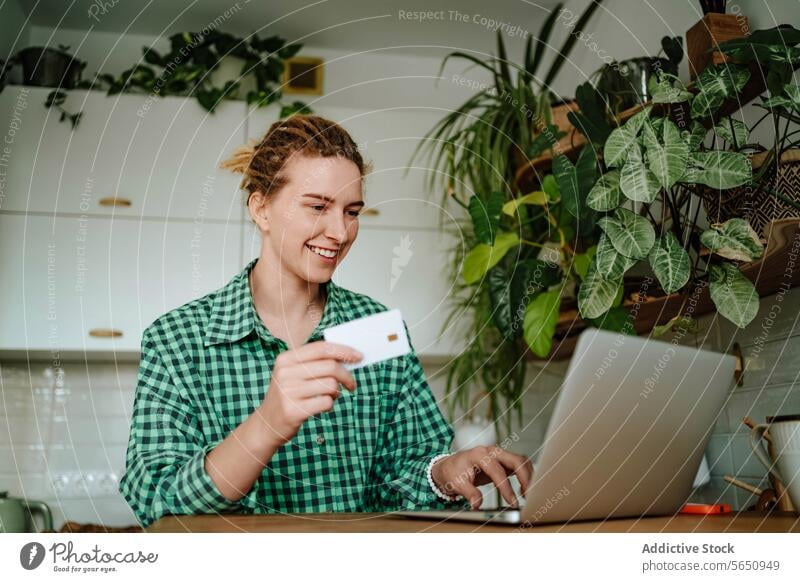 Young woman sitting with credit card near laptop at table in daylight at home purchase online smile shop happy glad female young positive device gadget shopper