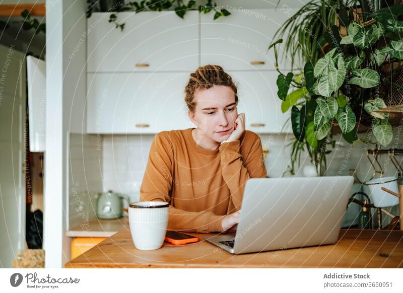 Young woman working on laptop at kitchen table with smartphone coffee mug in daylight thoughtful lean on hand cabinet recipe at home female young dreadlocks