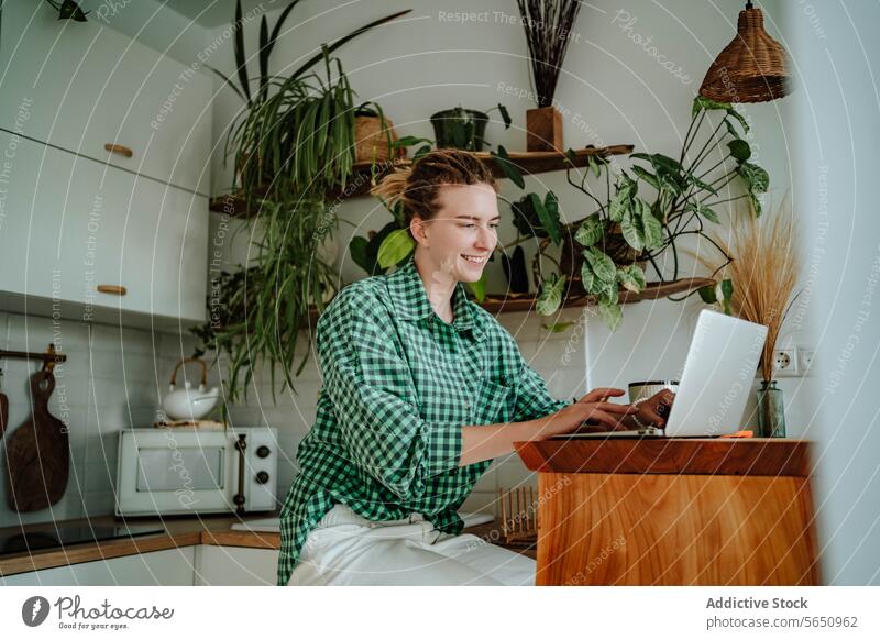 Happy young woman working on laptop at table in decorated kitchen at home freelance remote startup online female sit checkered shirt gadget device smile happy