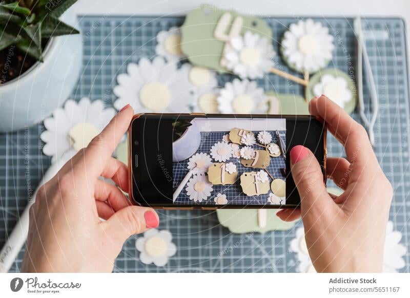 Woman framing a creative display of handmade paper tags and daisies through a smartphone screen photography crafting daisy flower DIY capture essence hobby