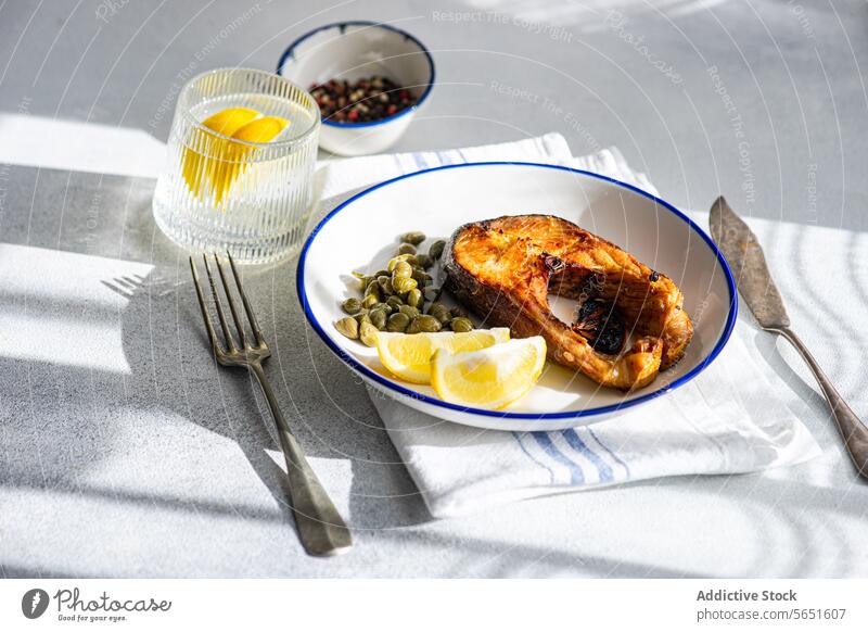 From above of Grilled Trout Steak on a Plate with capers and lemon placed on table near glass of water and cutlery trout steak grilled plate white blue rim