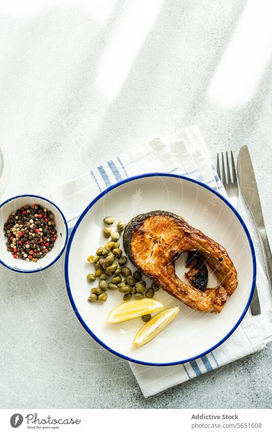 Top view of Grilled Trout Steak on a Plate with capers and lemon placed on table near cutlery and bowl trout steak grilled plate white blue rim garnish wedge