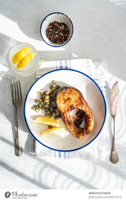 Top view of Grilled Trout Steak on a Plate with capers and lemon placed on table near glass of water and cutlery trout steak grilled plate white blue rim