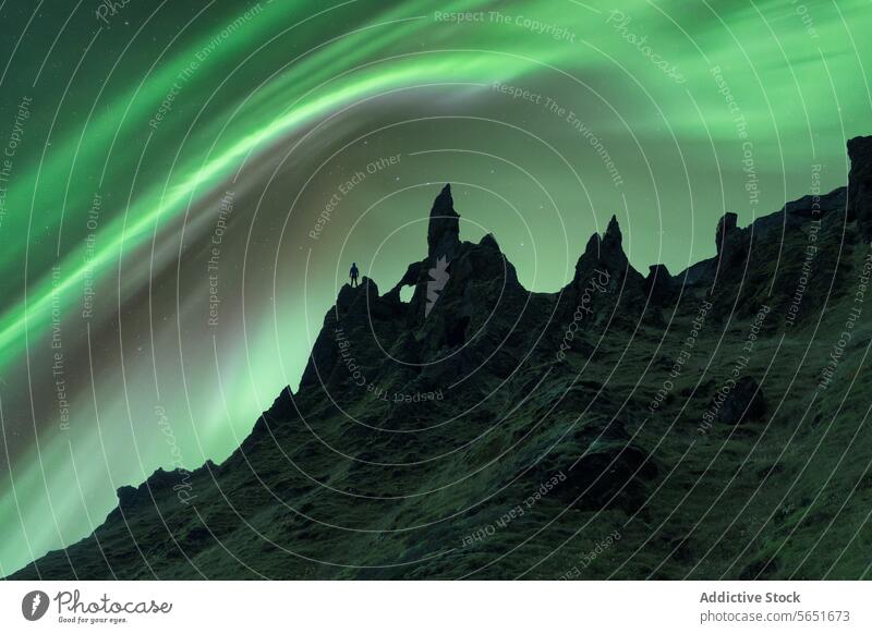 An unrecognizable person's silhouette stands out against jagged rock formations under the aurora borealis in Iceland. northern lights observer night green sky