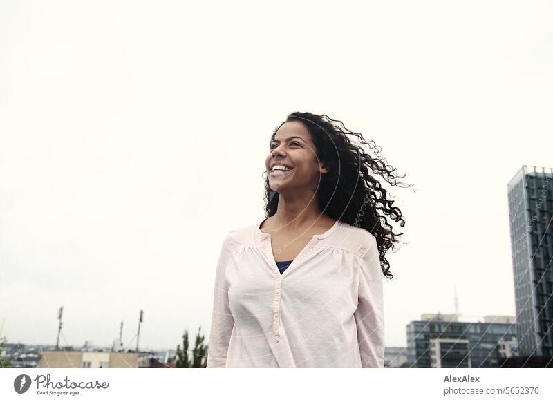 A young, beautiful woman stands on a rooftop in the city, looks up at the sky and laughs Woman youthful pretty Curl Black-haired Laughter Joy Beauty & Beauty