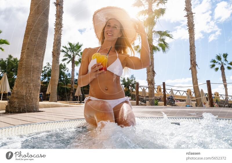 Woman in white bikini and straw hat enjoying a drink in a whirlpool, with palm trees around at a caribbean island hotel cocktail fun resort cheers woman sunny