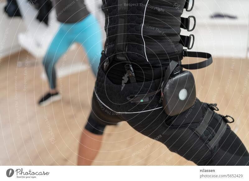 Two women performing squats in a gym, one wearing an EMS training suit with battery pack electro stimulation neuromuscular electrical personal training