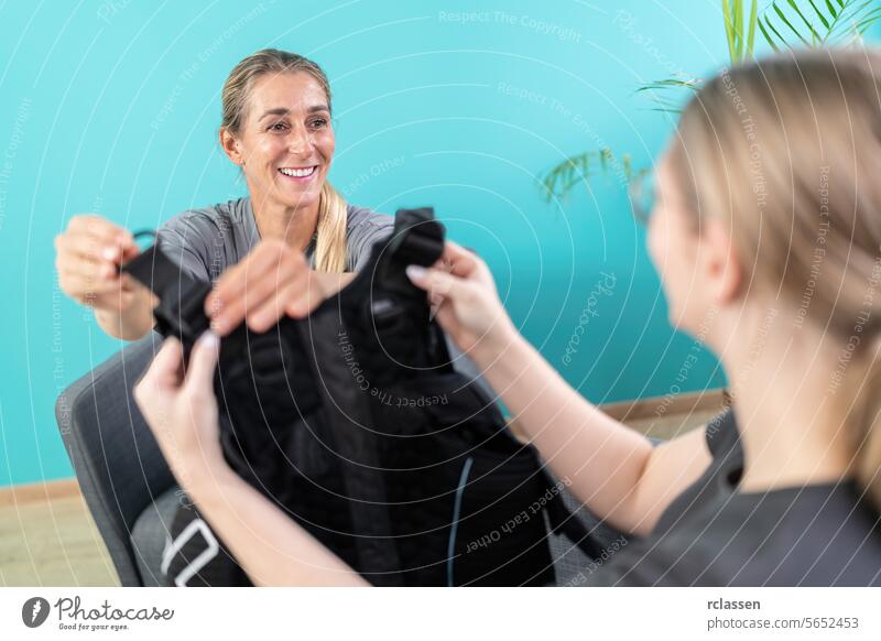 Trainer and client interacting with an EMS training vest in a bright room with a plant in background at a EMS - Stuido black vest handing over