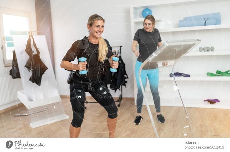 Woman with EMS training suit lifting dumbbells while trainer supervises with a tablet computer in a gym electro stimulation neuromuscular electrical