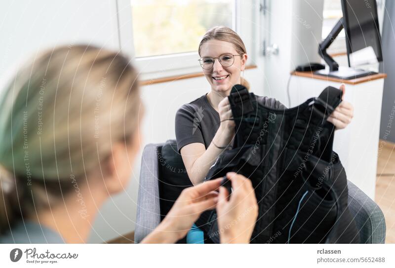 woman holding an EMS training vest explaining by talking to a trainer in a consultation room in a EMS - Studio ems training vest conversation armchairs indoor