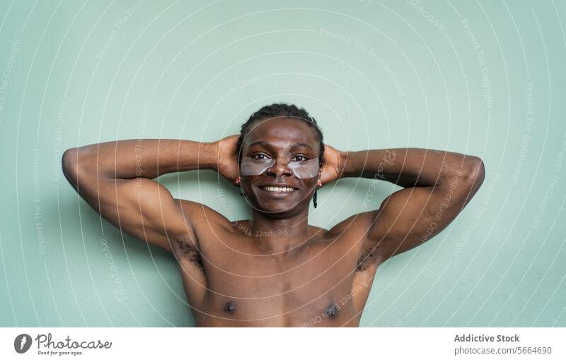 A joyful black man with under eye patches smiles at the camera hands behind his head against a mint green wall skincare under-eye patch happiness earring