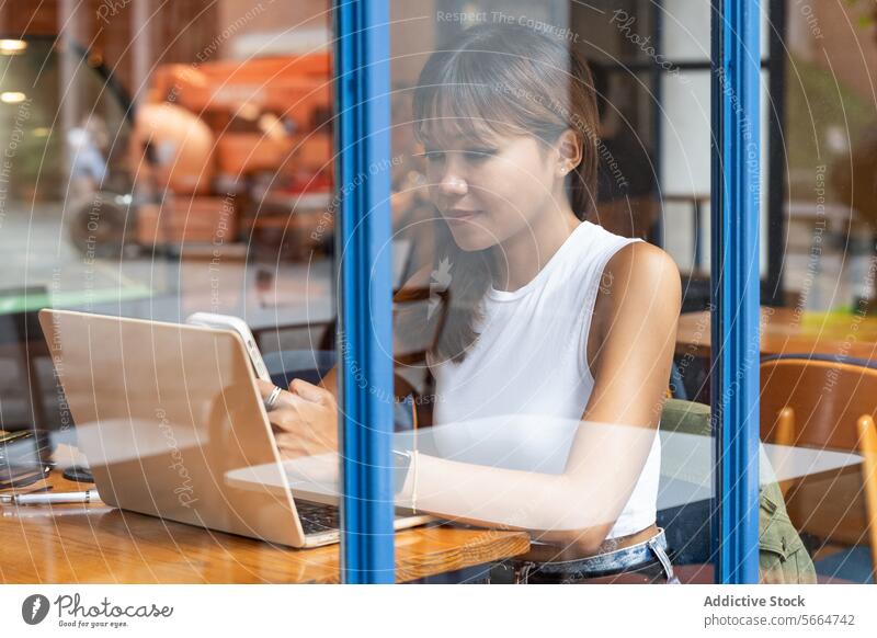 Through window Asian woman focused on her smartphone and tablet in a cafe during remote work in Chiang Mai, Thailand dedication mobile phone technology computer