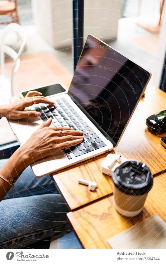 Cropped unrecognizable woman's hands typing on a laptop in a cafe, with a smartphone, earphones, and a coffee cup on the table during remote work in Chiang Mai, Thailand