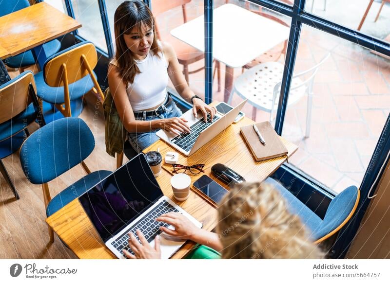 From above Asian woman in a white top working on her laptop in a cafe with a city view, engaging in remote work in Chiang Mai, Thailand, with a partially visible colleague beside herdigitak