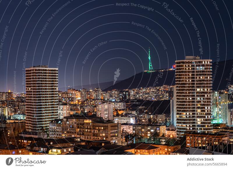 An urban night scape of Tbilisi showcasing the city's architecture with the TV tower illuminated on a hill in the background, under a dark blue sky lights