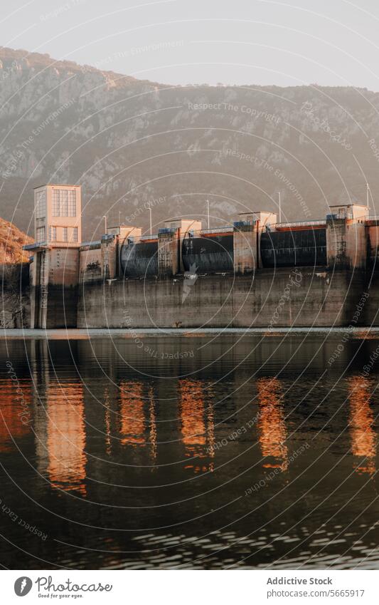 Warm sunset light bathes a dam creating a mirrored reflection on the still water surface against a backdrop of mountains warm infrastructure hydroelectric