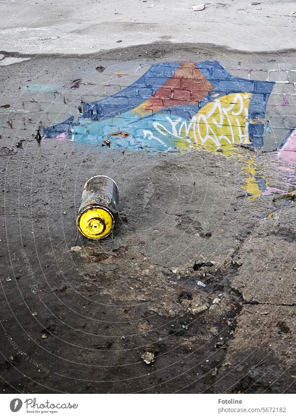 An empty spray can lies on the floor of a lost place. Unfortunately, a street artist left it there. Part of his artwork is reflected in the puddle behind it.